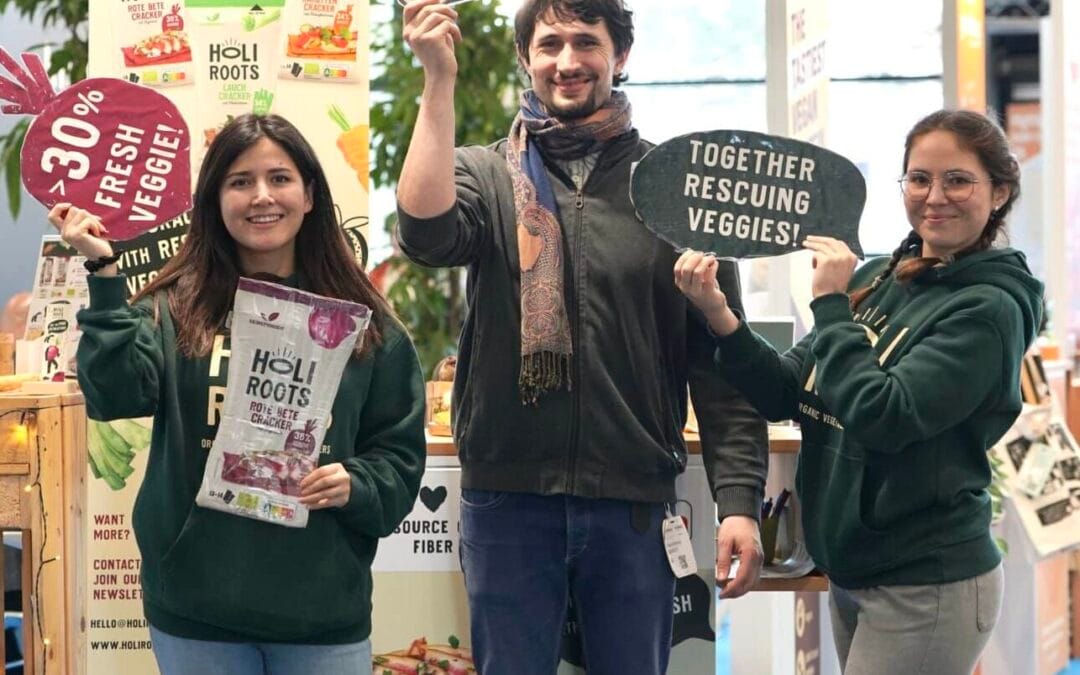 Holiroots Team hält Schilder mit Benefits der Biocracker in die Höhe: mehr als 30% Gemüse, Nutriscore A, Zusammen Gemüse Retten!